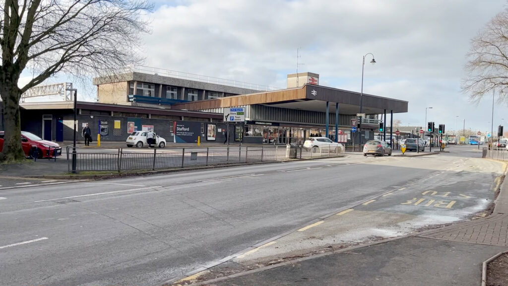 The facade of Stafford railway station, pictured from across the main road. The low, concrete building has a concrete roof that extends over the entrance.
