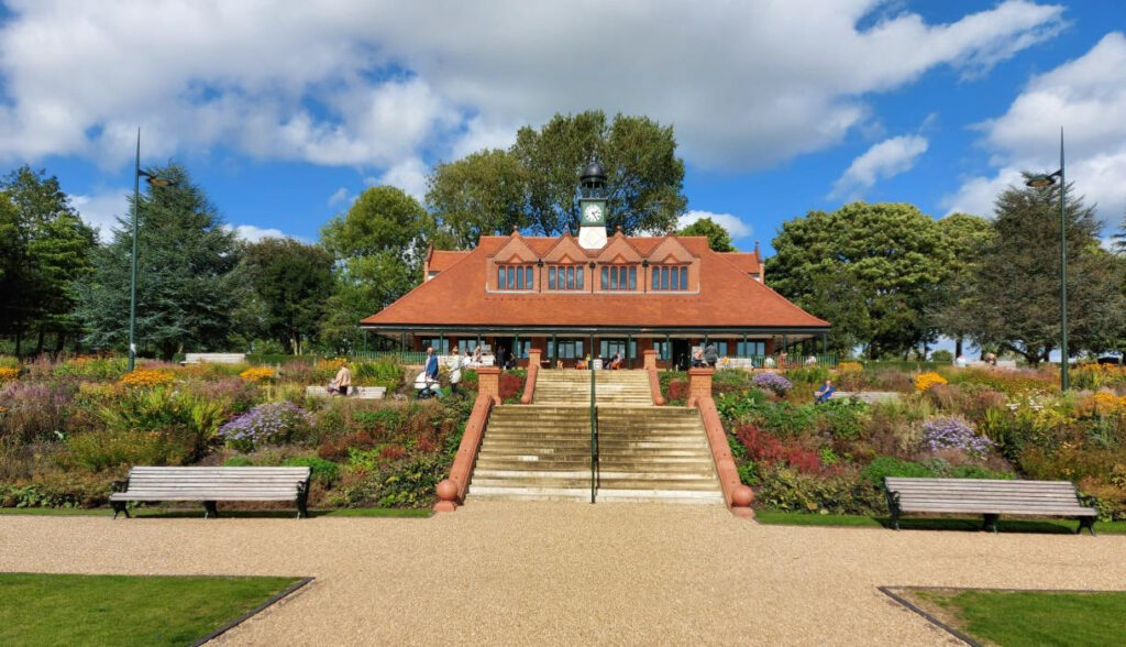 An image of Hanley Park, a large, gravelled park space with well maintained flower beds and a large building with clock tower.